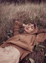Woman wearing a brown hooded top lying in tall dry grass, photographed outdoors in natural daylight by Nicole Bentley.