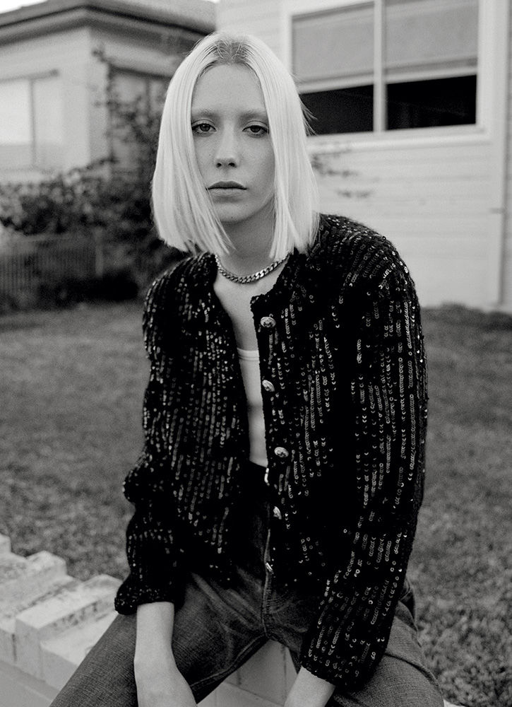 Monochrome portrait of a stylish blonde woman with a sequined jacket sitting outdoors against a house, with a serious facial expression.