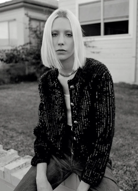Monochrome portrait of a stylish blonde woman with a sequined jacket sitting outdoors against a house, with a serious facial expression.