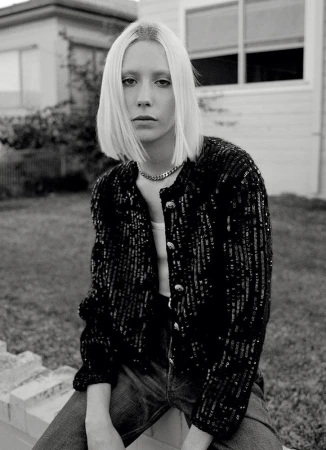 Monochrome portrait of a stylish blonde woman with a sequined jacket sitting outdoors against a house, with a serious facial expression.