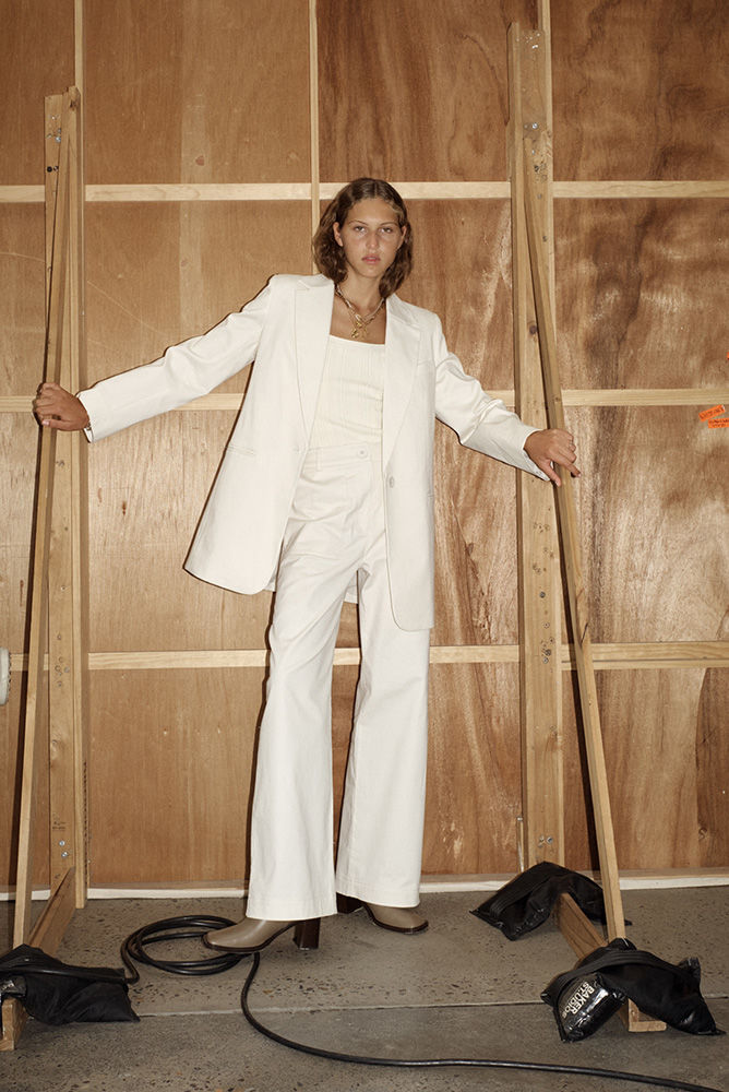A woman in a white blazer and pants stands against a wooden backdrop, striking a confident pose in a fashion-forward editorial shot.