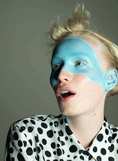 A young woman with pale skin and blonde hair wears a black and white polka-dot shirt and blue face paint in a studio setting.