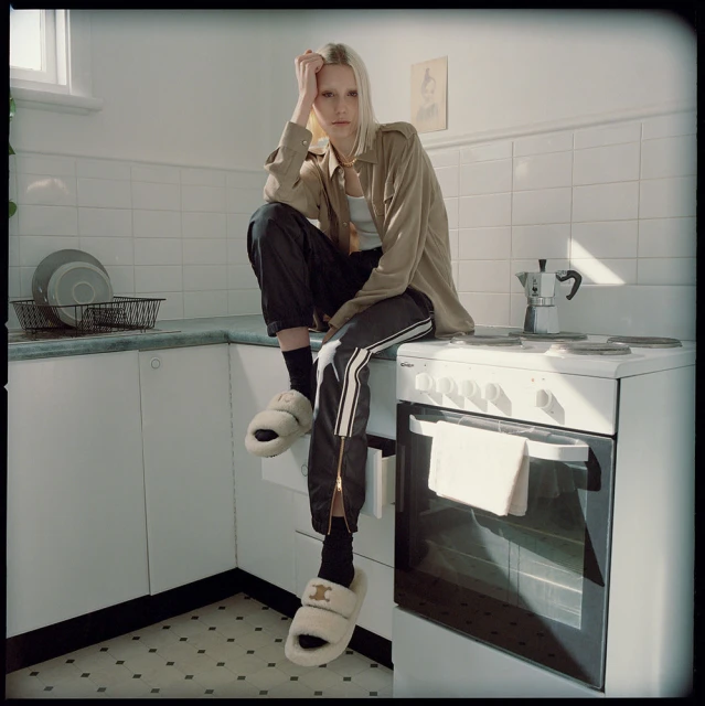 Young adult woman with blonde hair poses indoors in kitchen, wearing a jacket, pants, and fuzzy slippers. Natural light.