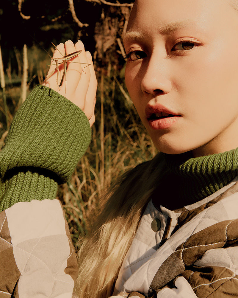 Close-up of a blonde female model posing with a stick insect on her hand outdoors with natural lighting and a quilted jacket.
