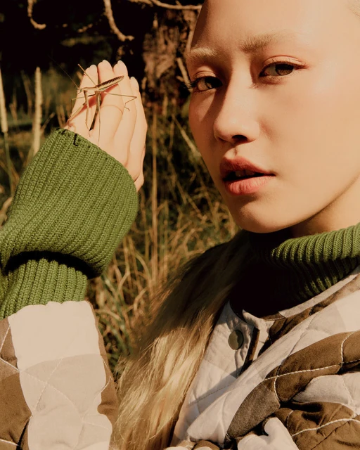 Close-up of a blonde female model posing with a stick insect on her hand outdoors with natural lighting and a quilted jacket.