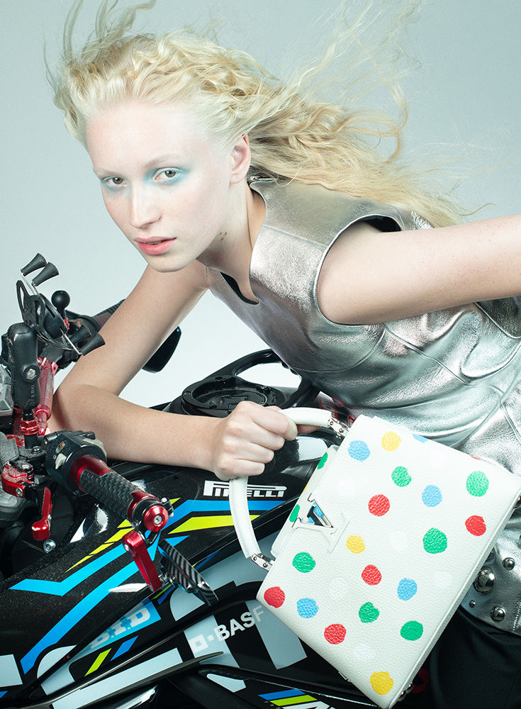 Fashion shot of a fair-skinned woman with white-blonde hair in a metallic silver dress, posing with a motorcycle and designer handbag.