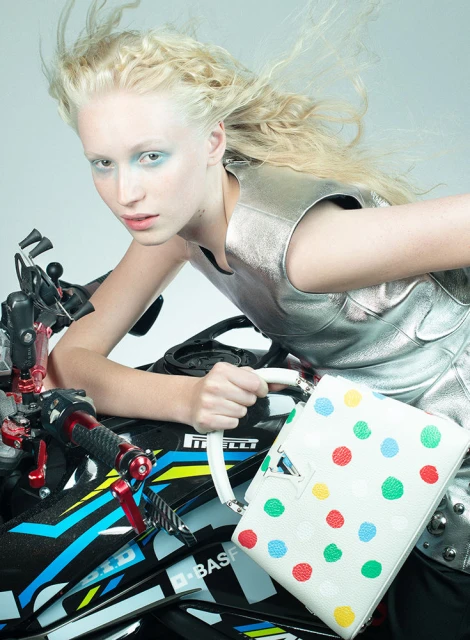 Fashion shot of a fair-skinned woman with white-blonde hair in a metallic silver dress, posing with a motorcycle and designer handbag.