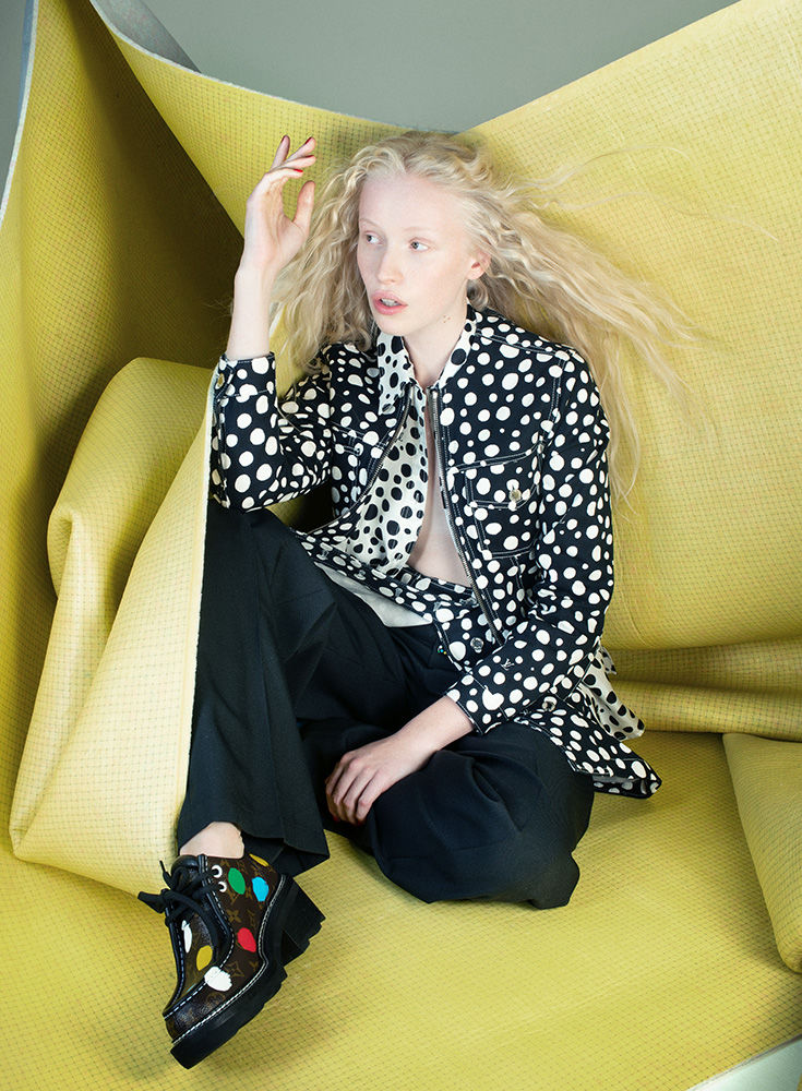 Stylish albino woman in a polka dot jacket poses inside a draped yellow fabric setting, wearing black pants and unique platform shoes.