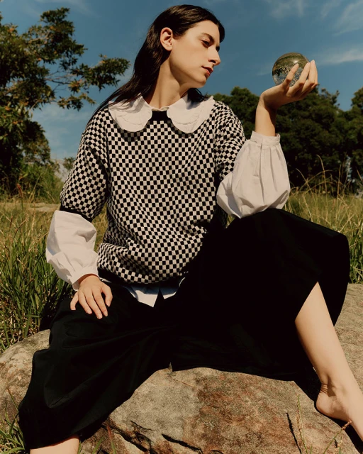Woman in black and white checker sweater poses with a crystal ball in an outdoor setting with trees, blue sky, and golden grass.
