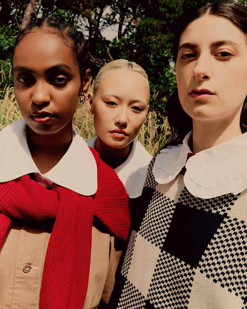 Three young women wearing preppy fashion stand closely together in an outdoor setting with trees and tall grass in the background.