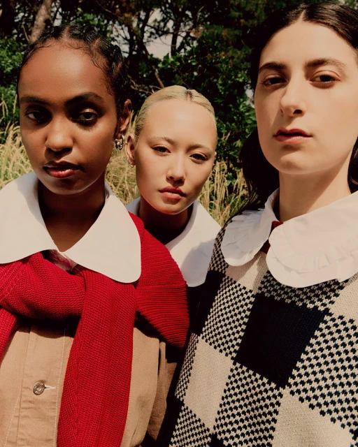 Three young women wearing preppy fashion stand closely together in an outdoor setting with trees and tall grass in the background.