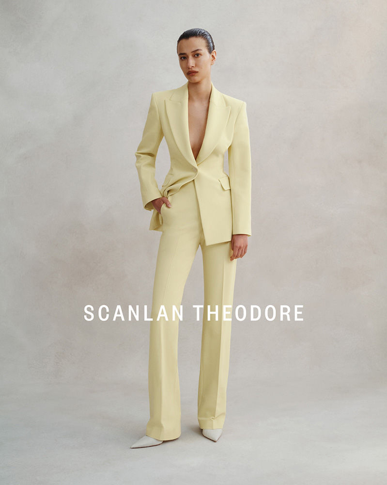 A woman models a light-yellow pantsuit with a blazer and flared pants, posing in front of a neutral-toned background in a studio setting.