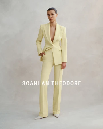 Model wearing a pale yellow tailored pantsuit with white pointed-toe shoes against a neutral textured studio background