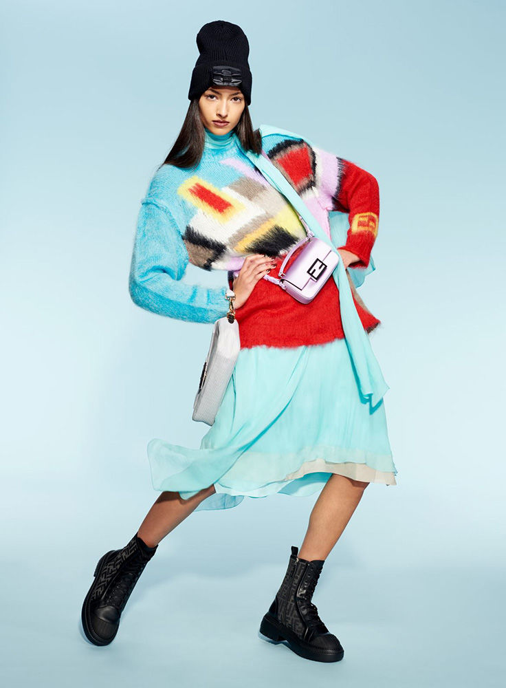 Fashionable young woman in a colorful sweater, mini skirt, black beanie, and boots, carrying a small pink purse on a blue background.