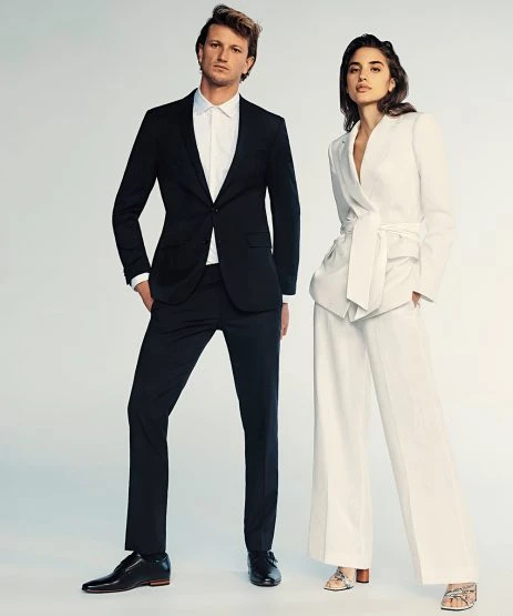 Elegant fashion shot of a man in a black suit and a woman in a white suit standing in a bright photo studio setting.