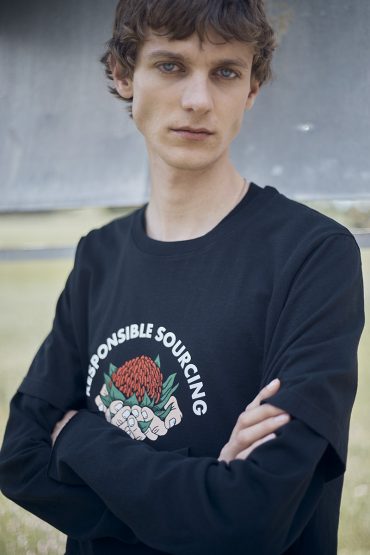 A young adult male with curly brown hair poses in a black long-sleeved shirt with a responsible sourcing graphic against a neutral background.