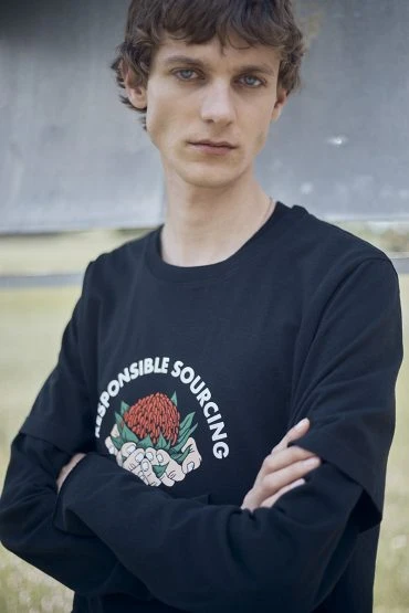 A young adult male with curly brown hair poses in a black long-sleeved shirt with a responsible sourcing graphic against a neutral background.