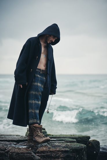 Adult male wearing a hooded coat and patterned trousers stands on a rocky outcrop by the sea in an outdoor setting, overcast weather.