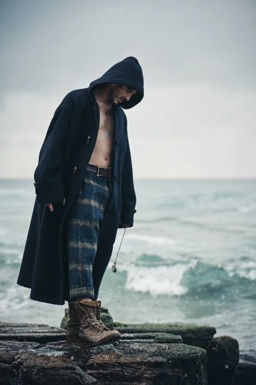 Adult male wearing a hooded coat and patterned trousers stands on a rocky outcrop by the sea in an outdoor setting, overcast weather.