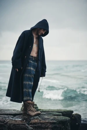 Adult male wearing a hooded coat and patterned trousers stands on a rocky outcrop by the sea in an outdoor setting, overcast weather.
