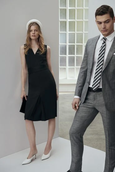 Two fashion models pose in a photo studio. The woman wears a black dress and white shoes and the man wears a grey suit.