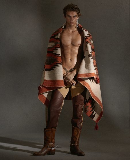 A muscular adult male model poses indoors wearing chaps and cowboy boots, draped in a Southwestern-patterned blanket.