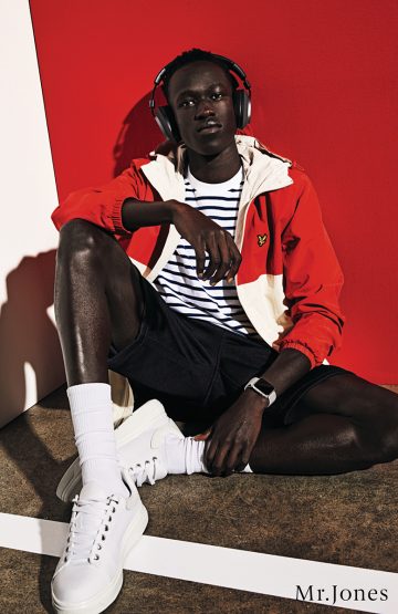 Dark-skinned man in headphones, jacket, and shorts poses on a red-and-white set, showcasing white sneakers and socks in a fashion context.