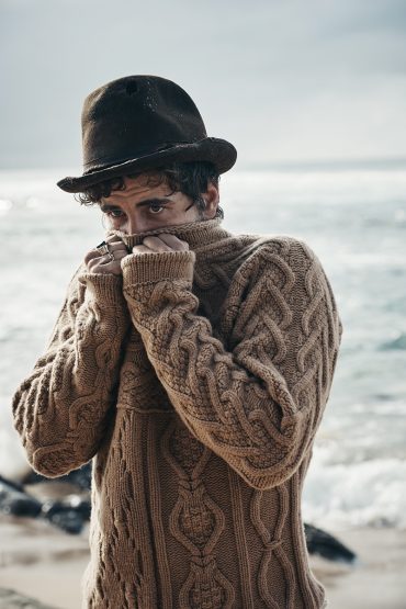 Man in a brown knit sweater and hat stands on a sandy beach with the ocean in the background, face partially covered by the sweater.