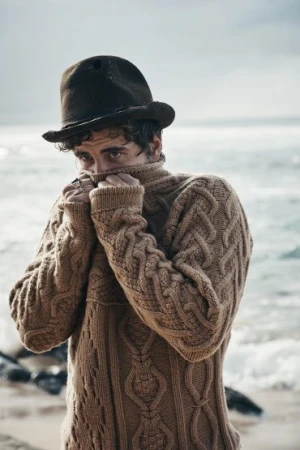 Man wearing a brown cable knit sweater and dark felt fedora pulling the collar over his face on a beach