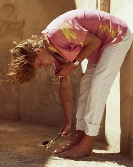 Man with blond hair bends over, barefoot, wearing a pink shirt and white pants, picking up keys off the floor in a tan room.