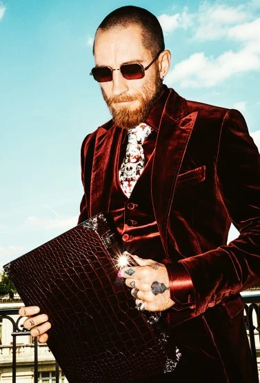 Stylish man in a maroon velvet suit with sunglasses holding a leather portfolio on a balcony against a bright blue sky background.