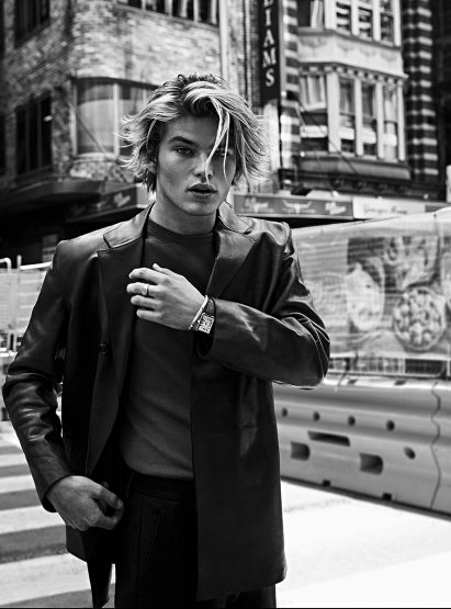 Black and white portrait of a male model with blond hair wearing a blazer in an urban setting, standing on a crosswalk.