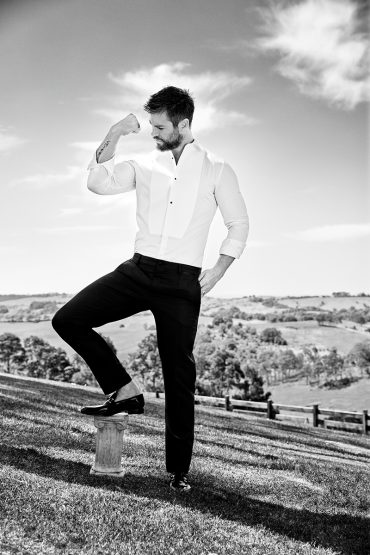 Monochrome portrait of a man in formal wear flexing outdoors with a blue sky and grassy hills in the background on a sunny day.