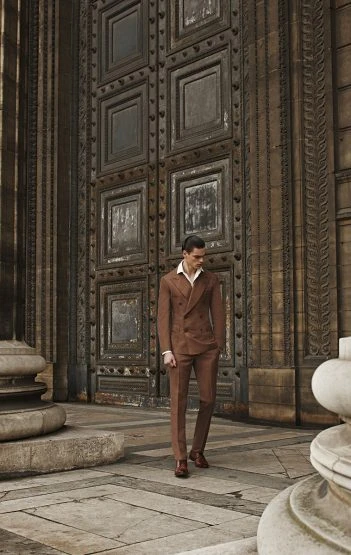 Man in brown suit standing in front of large, ornate doorway. Full shot, fashion editorial, stone architecture, warm color grading.