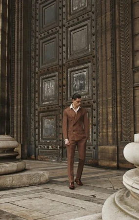 Man in brown suit standing in front of large, ornate doorway. Full shot, fashion editorial, stone architecture, warm color grading.