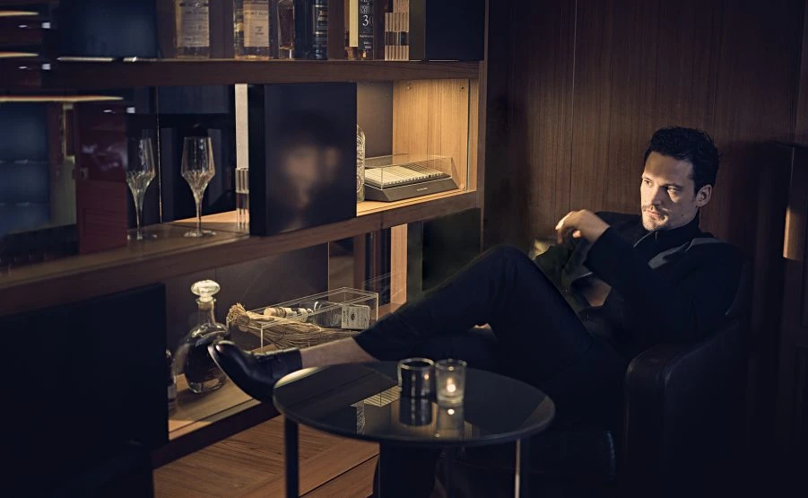 Adult man in black turtleneck reclines in dark armchair in wood-paneled bar; glasses, liquor bottles on lit shelves in the background.