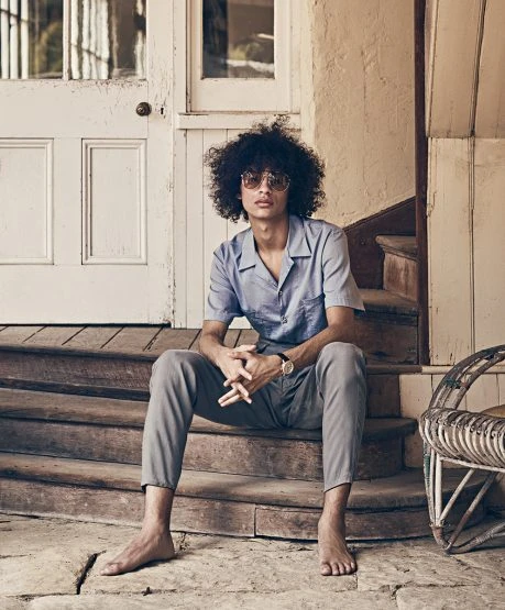 Young adult male with afro sits on wooden steps outdoors wearing a blue camp shirt, grey pants, and sunglasses in the daytime.