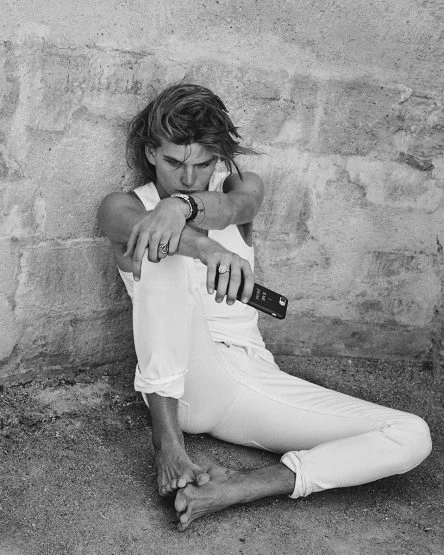 A young adult male with blond hair sits with knees up, holding a smartphone against a textured wall in a black-and-white fashion shot.