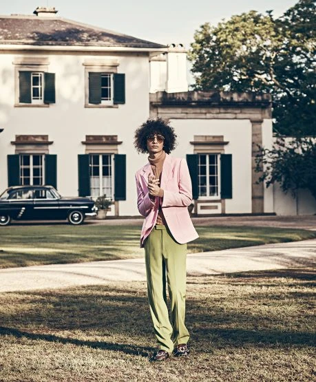 A man in a pink jacket and green pants stands on a lawn in front of a white building and a black car on a sunny day.