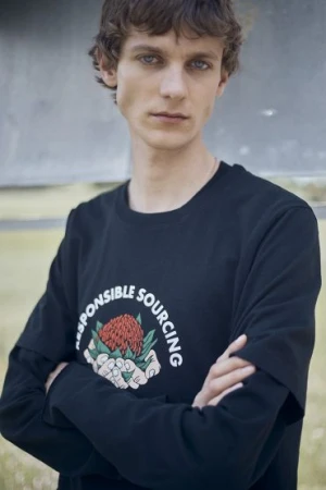 A young male model with brown curly hair wearing a black long-sleeve t-shirt with a graphic print, posed outdoors.