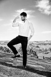 Man wearing a tuxedo shirt and dark trousers flexing his bicep outdoors on a grassy hill under a cloudy sky.