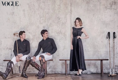 Two male polo players in black shirts and white trousers seated on a bench next to a standing woman in a black gown.
