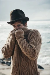 Man wearing a brown cable knit sweater and dark felt fedora pulling the collar over his face on a beach