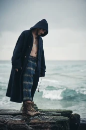 Man wearing a dark hooded coat over bare chest and plaid trousers standing on rocky shore by the ocean