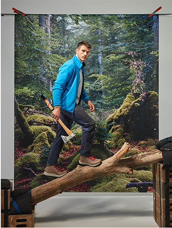 A man in a blue jacket is walking on a log with an axe against a forest backdrop in a studio setting.