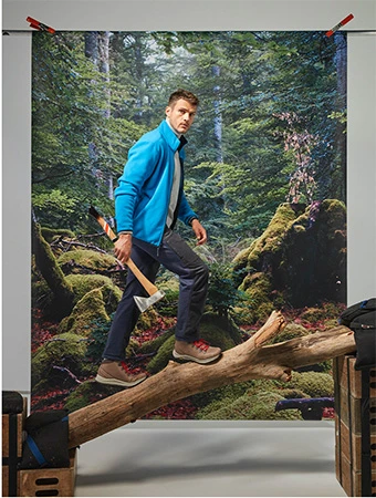 A man in a blue jacket is walking on a log with an axe against a forest backdrop in a studio setting.