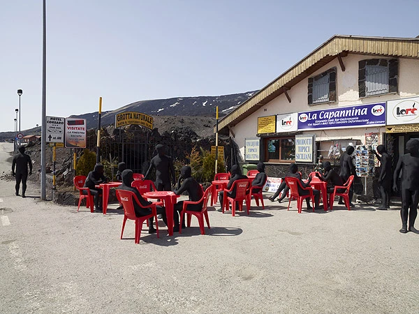 Art installation featuring figures in black bodysuits seated and standing around red tables and chairs outside a building on a sunny day.