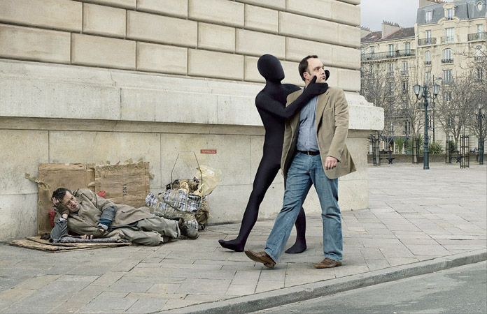 A man in a suit is being grabbed by a person in a black suit while another man lies on the ground in a city square.