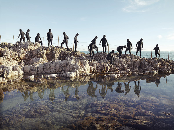 Group of people in wetsuits on a rocky shoreline with reflections in the water, against a blue sky, captured in a sunny outdoor shot.