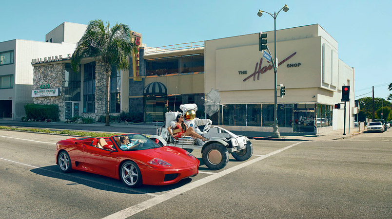 Red convertible car and moon rover with a woman and astronaut, on a city street with vintage buildings and traffic lights in daylight.